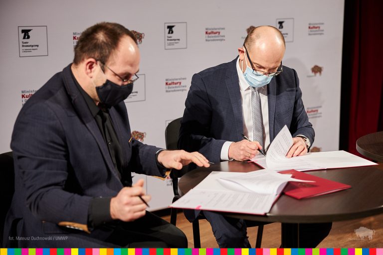 Dyrektor teatru, który znajduje się po lewej stronie i marszałek, po prawej stronie podpisują umowę. Za nimi znajduje się ścianka, na której jest napisane Teatr Dramatyczny