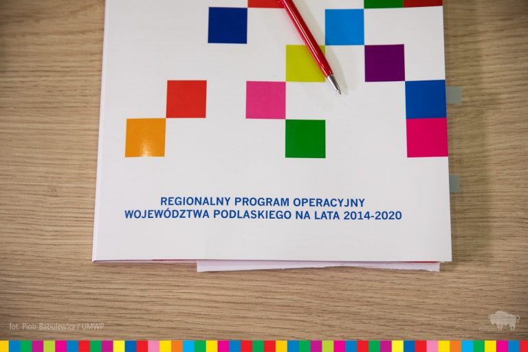 Na stoliku leży papierowa teczka, biała w niebiesko - fioletowo - różowo - zielono - pomarańczowe kwadraty