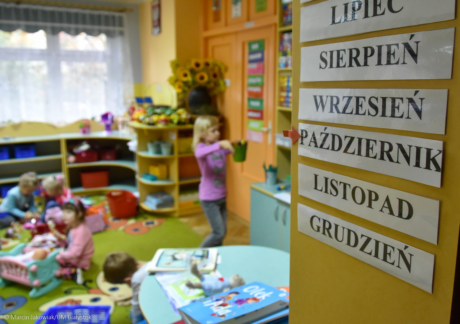 Dzieci bawią się w sali w przedszkolu. Po prawej stronie zdjęcia na ścianie przyczepione są kartoniki z nazwami miesięcy od lipca do grudnia