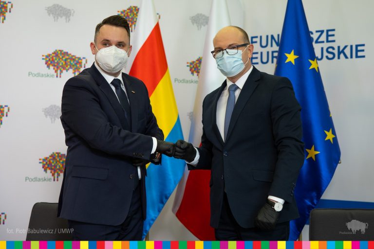 Dwóch mężczyzn; z lewej strony zastępca prezydenta Białegostoku, z lewej marszałek przybijają tzw. żółwika. Obaj są w ciemnych garniturach. Stoją na tle flagi Polski, Unii Europejskiej oraz województwa podlaskiego