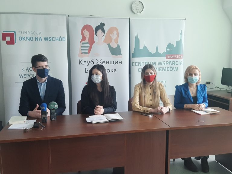 Przy brązowym stole siedzę cztery osoby, od lewej strony mężczyzna, następnie kobiety, Za nimi stoją banery promocyjne
