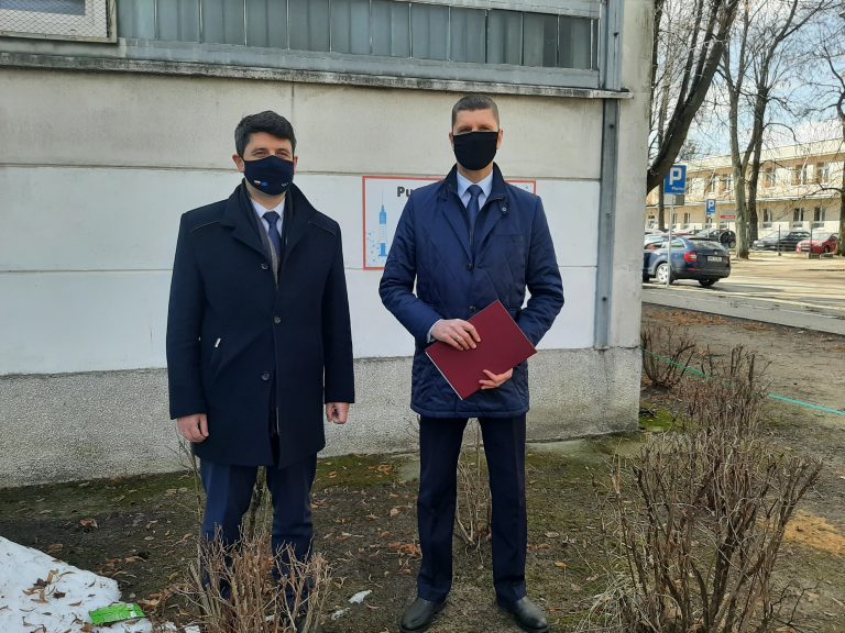 Poseł Mariusz Gromko, Dariusz Piontkowski stoją na tle betonowej ściany. Mężczyźni są ubrani w granatowe garnitury i płaszcze