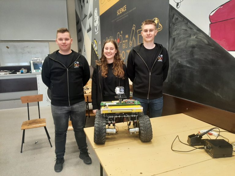 Trzy młode osoby, po bokach chłopaki, w środku dziewczyna stoją w pracowni. Przed nimi na stole stoi robot, które samodzielnie stworzyli