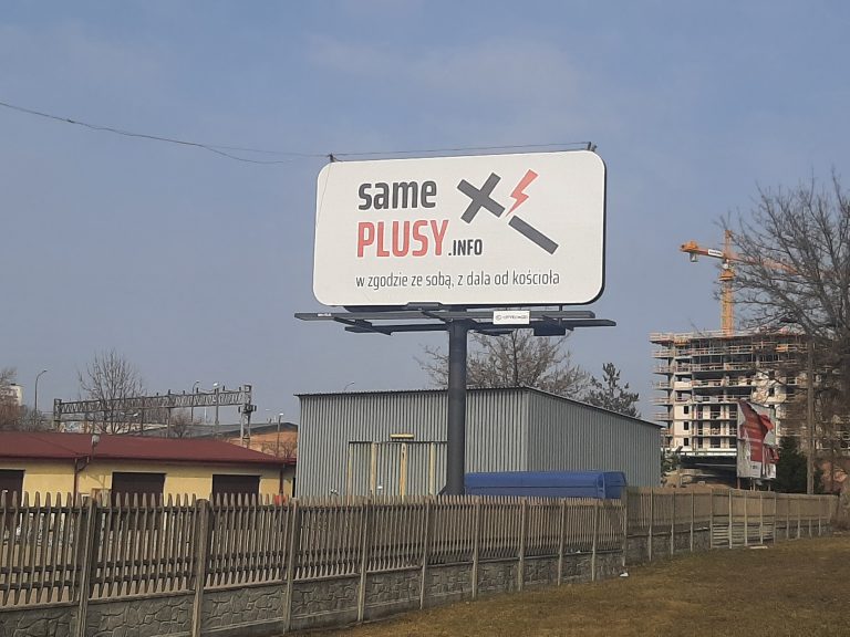 Bilbord z napisem same plusy, na który jest również pęknięty krzyż. Za bilbordem widoczny jest budujący się blok i dźwig budowlany