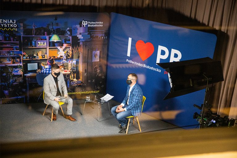 Studio telewizyjne, z góry widoczni są dwaj mężczyźni, którzy ze sobą rozmawiają. Po prawej stronie jest niebieska ścianka Politechniki Białostockiej z hasłem: "I love PB"