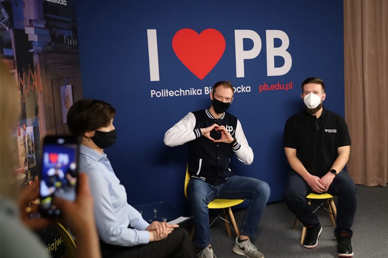 Dwóch studentów siedzi na krzesłach na tle ścianki, na której jest napis: "I love PB". Chłopak siedzący po lewej strony układa dłonie w kształt serca, również z lewej strony siedzi kobieta, która ma na sobie jasną koszulę oraz na krótko ścięte włosy. Tym trzem osobom, dziewczyna robi zdjęcia telefonem. Kobieta znajduje się w lewym rogu