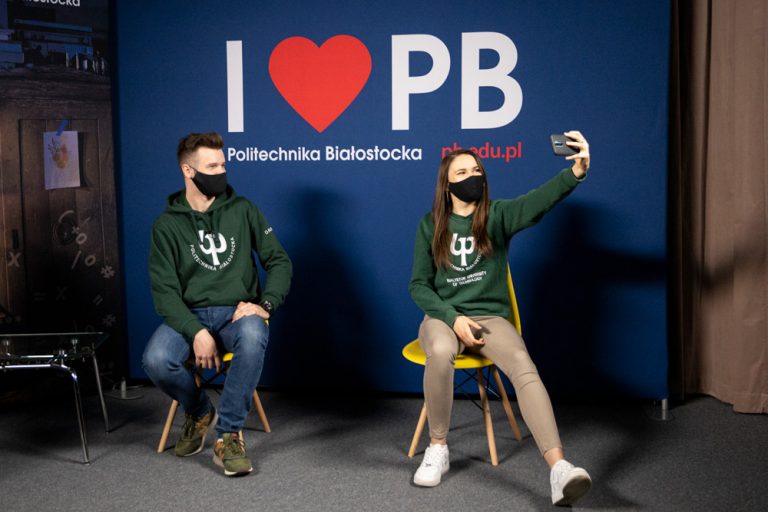 Student i studentka siedzą na krzesłach na tle niebieskiej ścianki z napisem "I love PB". Ubrani są w zielone bluzy z białym logiem PB. Dziewczyna, która znajduje się po prawej stronie robi selfie telefonem komórkowym