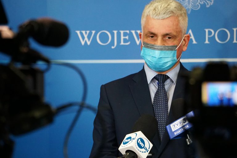 Wojewoda podlaski jest w ciemnym garniturze stoi na tle niebieskiej ścianki. Kadr pochodzi z konferencji prasowej, przed wojewoda są mikrofony oraz kamery
