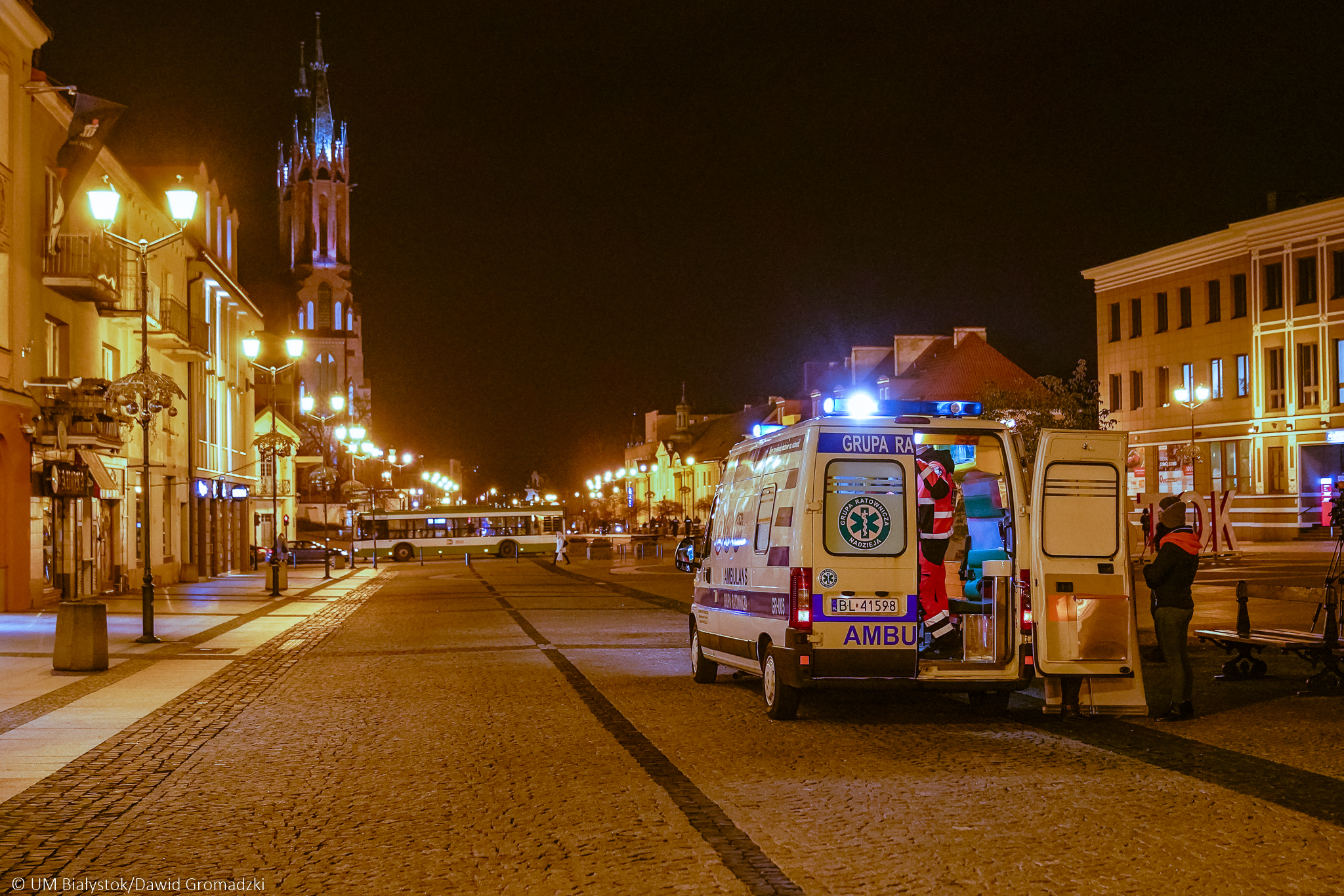 Rynek Kościuszki w Białymstoku. nocą. Miasto jest oświetlone, w oddali widać wieżę katedry, przejeżdżający autobus, po prawej stronie stoi ambulans