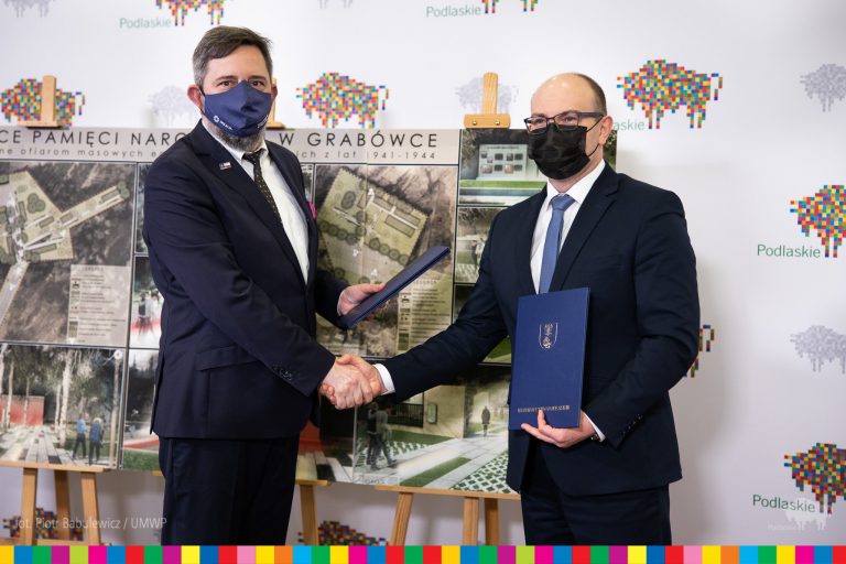 Dwóch mężczyzn w ciemnych garniturach i maseczkach ściska sobie dłonie. Stoją na tle plansz z projektami Miejsca Pamięci Narodowej w Grabówce. Za stojakami z planszami jest ścianka województwa podlaskiego, biała w pikselowo - kolorowe żubry