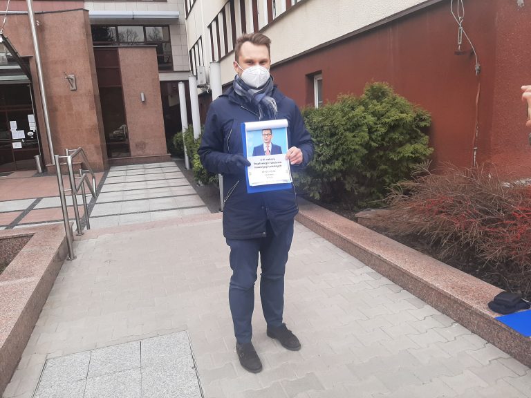 Mężczyzna stoi na tle Podlaskiego Urzędu Wojewódzkiego przy ul. Mickiewicza w Białymstoku. W rękach trzyma wydrukowaną podobiznę premiera Morawieckiego