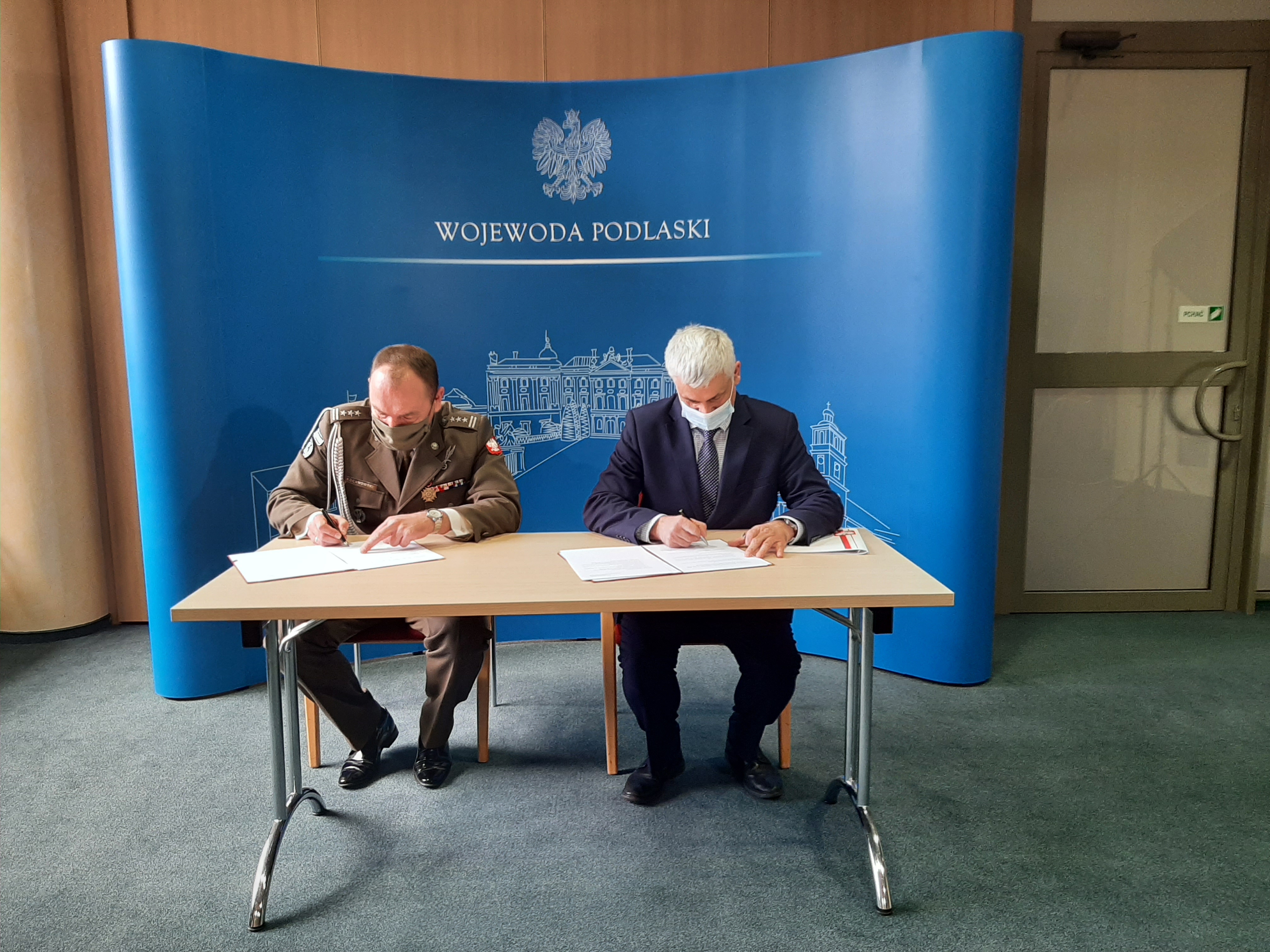 Dwóch mężczyzn siedzi przy stole, pochyleni podpisują umowę. Za nimi znajduje się niebieska ścianka Podlaskiego Urzędu Wojewódzkiego
