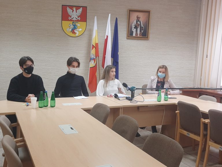 Przy stole siedzą cztery osoby, za nimi znajdują się flagi: Inii Europejskiej, Polski i Białegostoku. Na ścianie wisi herb miasta i obraz błogosławionego księdza Michała Sopoćki, patrona miasta
