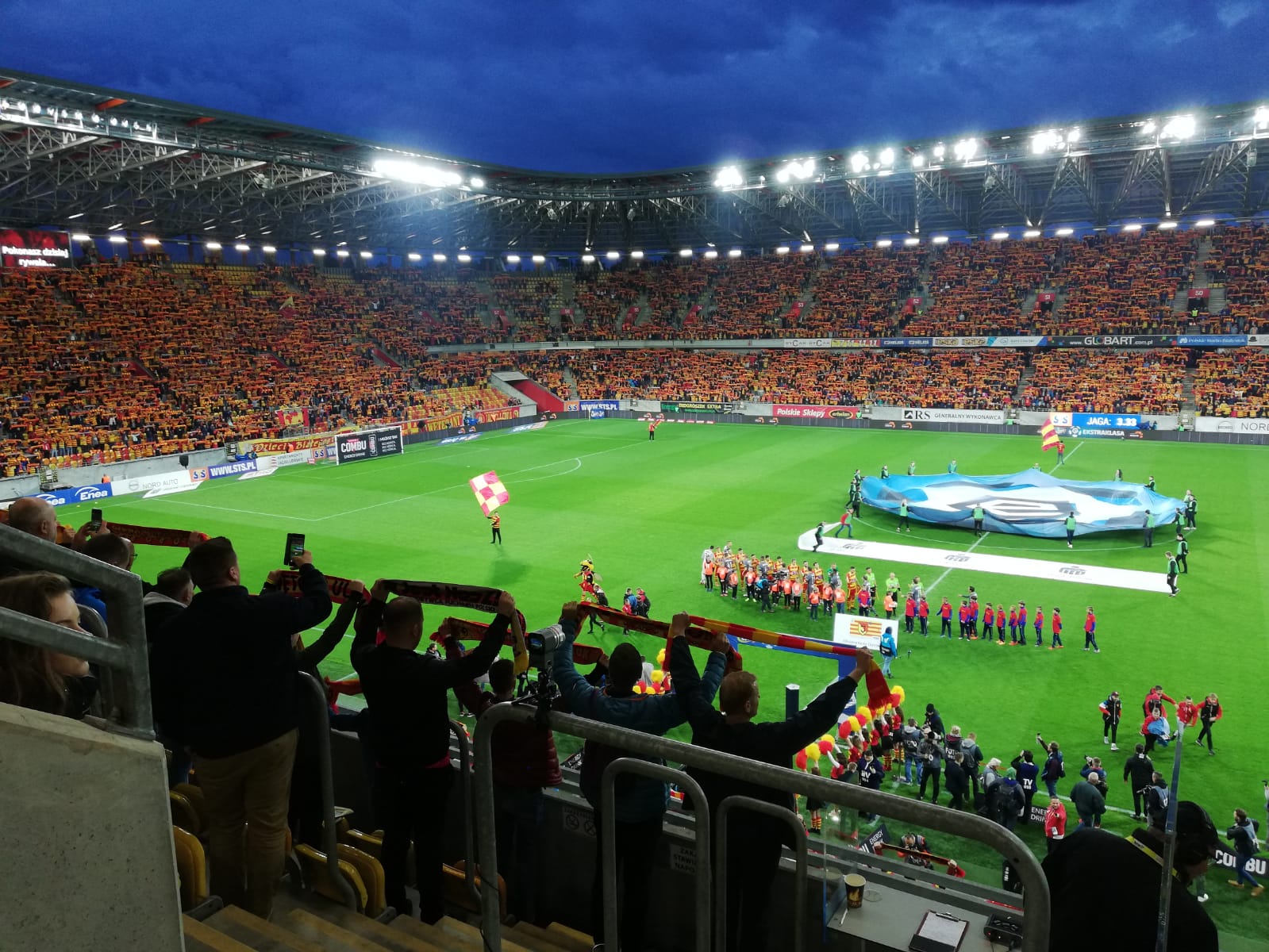 Stadion Miejski w Białymstoku, obiekt jest podświetlony. Na trybunach są kibice, którzy trzymają w górze klubowe szaliki
