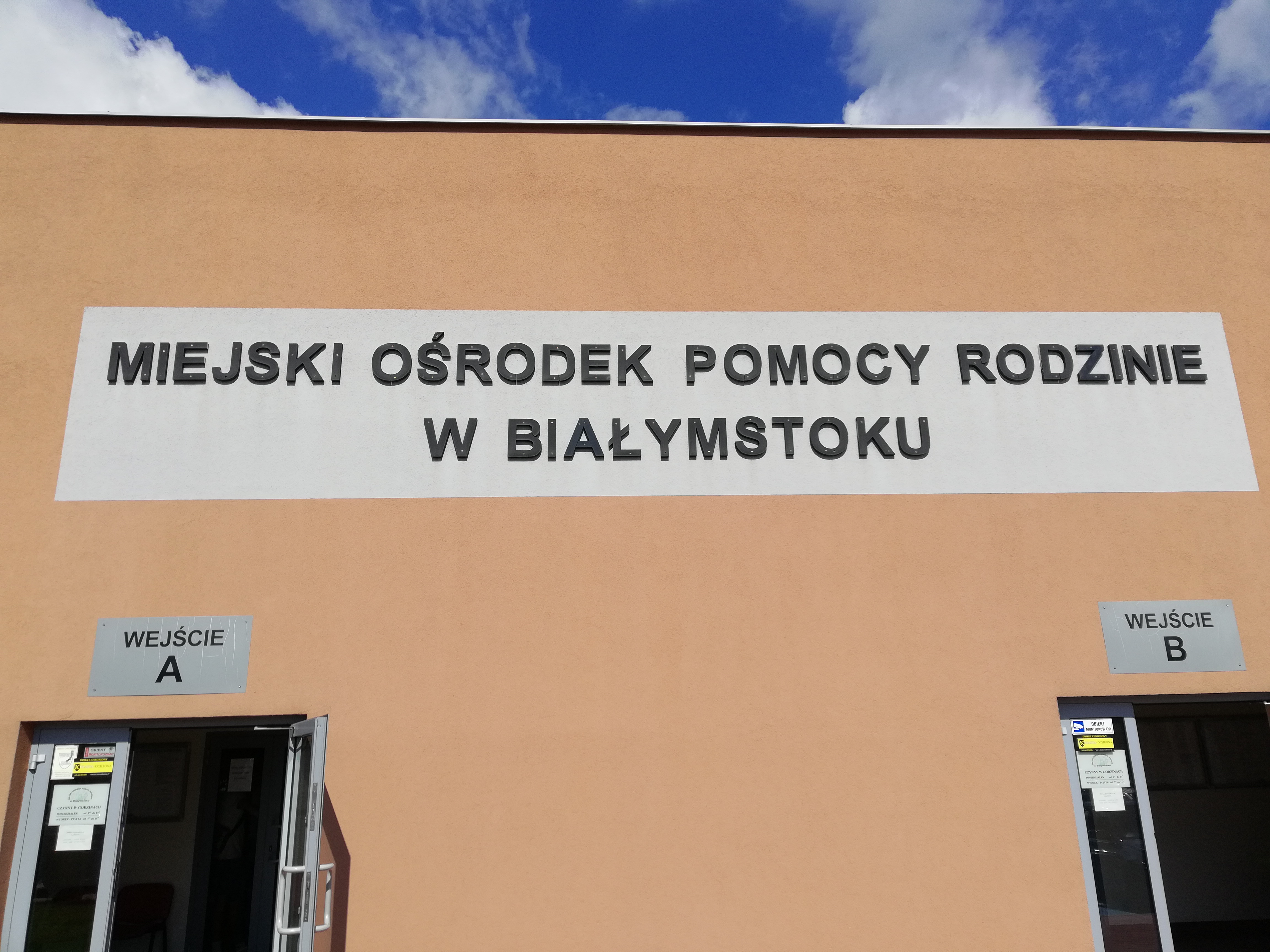 Fragment ściany budynku w kolorze jasnej cegły., na której na szarym pasku znajduje się czarny napis: Miejski Ośrodek Pomocy Rodzinie w Białymstoku