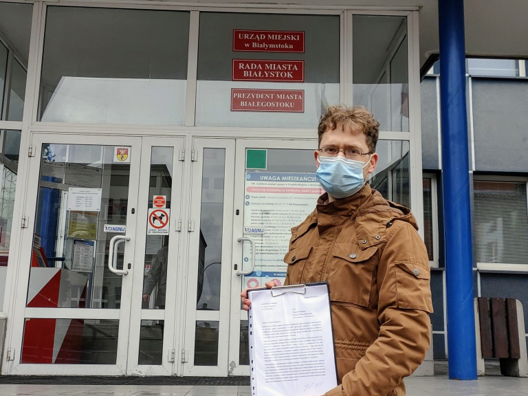 Mężczyzna w maseczce, który prezentuje petycję stoi na tle wejścia do Urzędu Miejskiego przy ul. Słonimskiej w Białymstoku