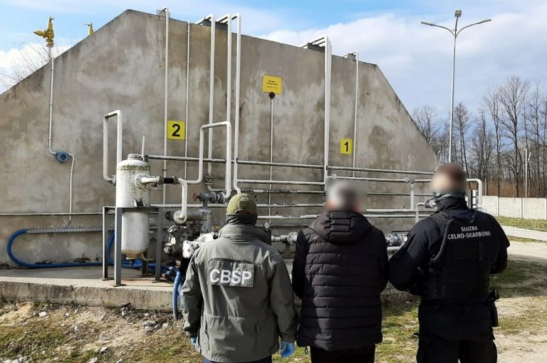 Trzy osoby przed aparaturą przesyłową gazu, z lewej stoi funkcjonariusz CBŚP z prawej funkcjonariusz Służby Celno-Skarbowej