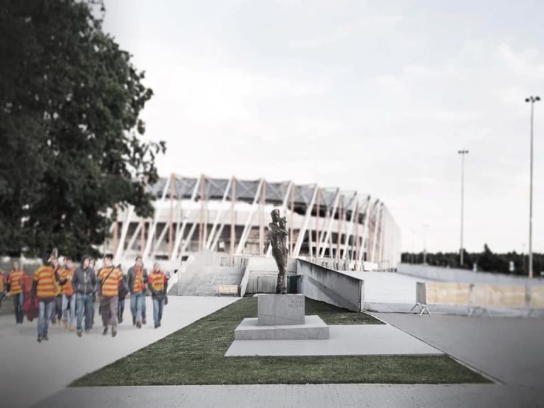 W oddali widać Stadion Miejski w Białymstoku.. Z lewej strony idą kibice w koszulkach w żółto - czerwone paski. Na środku zdjęcia znajduje się pomnik 42. Pułku Piechoty