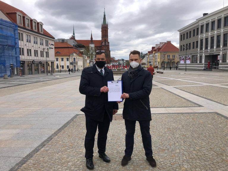 Dwóch posłów; po lewej stronie - Stefan Krajewski, z prawej Krzysztof Truskolaski trzymają wspólnie wniosek, który przygotowali. Mężczyźni stoją na Rynku Kościuszki w Białymstoku, za nimi widać Katedrę