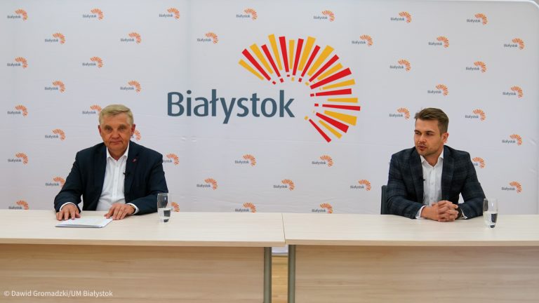 Dwóch mężczyzn siedzi przy stole na tle ścianki z napisem Białystok i z logiem wschodzącego słońca. Z lewej strony siedzi prezydent Białegostoku.