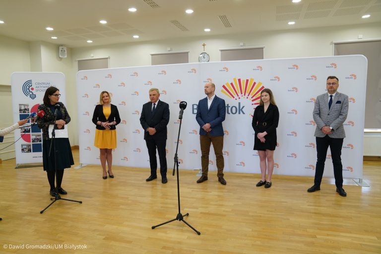 Na ściance z logo Białegostoku, którym jest wschodzące słońce stoi sześć osób