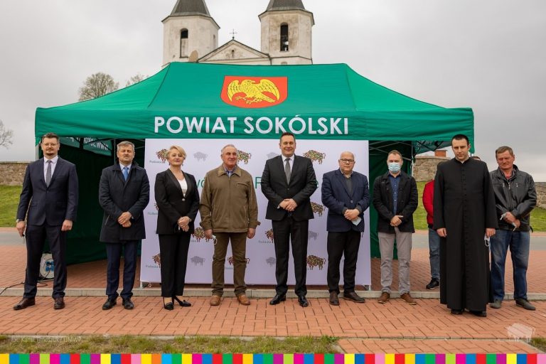 Ludzie stoją w rzędzie za nimi stoi zielony namiot z napisem Powiat Sokólski oraz kościół