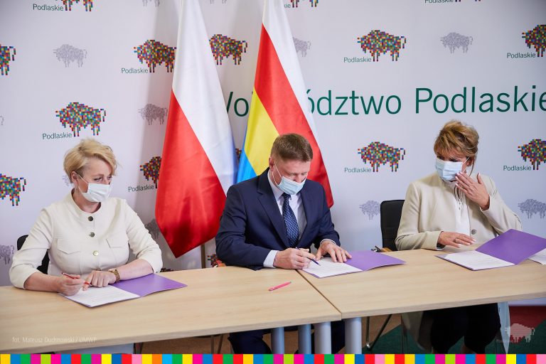 Przy stole konferencyjnym siedzą trzy osoby, podpisują porozumienie