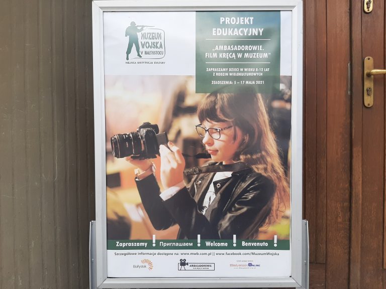 Przy drzwiach stoi plakat, na którym widoczna jest dziewczynka z aparatem na szyi i która patrzy w ekran urządzenia