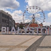 Okolice Rynku Kościuszki w Białymstoku, centralnie na placu ustawiony jest biały napis hasztag Białystok, za napisem widoczna jest karuzela typu diabelski młyn
