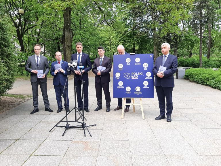W Parku Plany, w rzędzie stoją politycy Prawa i Sprawiedliwości. Między nimi stoi niebieski plakat