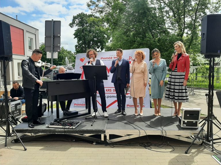 Na scenie stoją soliści i śpiewają. Z lewej strony mężczyzna gra na pianinie, a drugi na skrzypcach