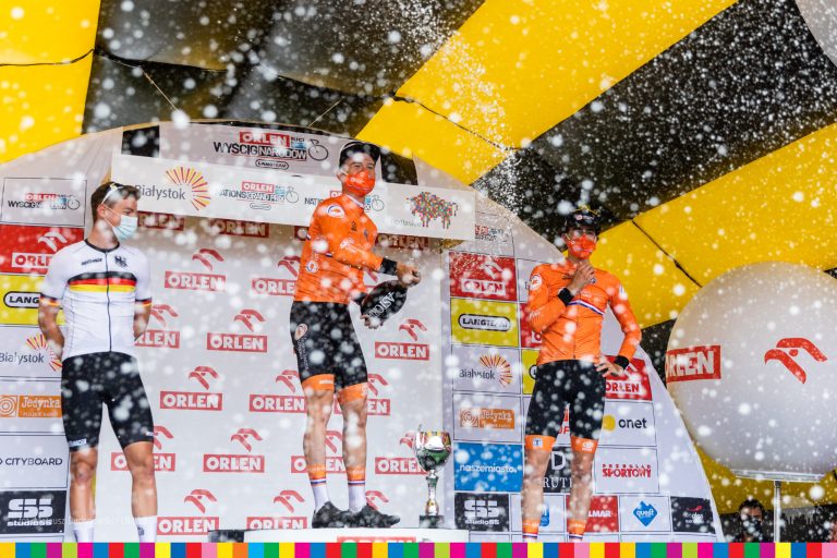 Trzej mężczyźni - kolarze stoją na podium. Zwycięzca, który stoi najwyżej na podium rozlewa szampana