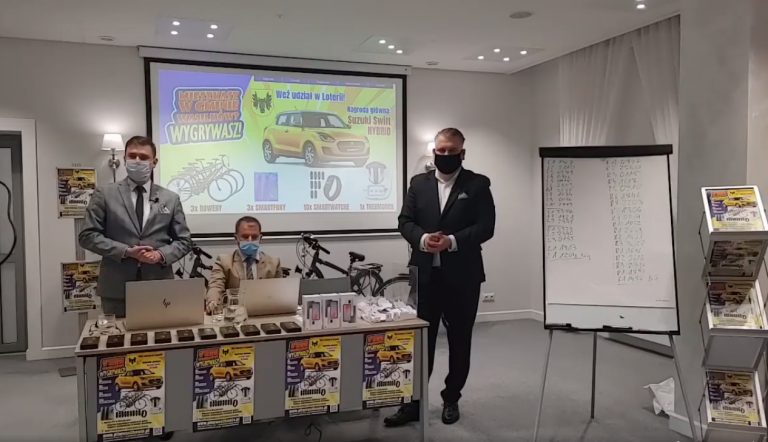 Sala konferencyjna, w której odbywa się losowanie nagród . Dwóch mężczyzn stoi przy stole, jeden siedzi pośrodku. Za nimi znajduje się rzutnik, na którym wyświetlane są nagrody, które można zdobyć w loterii