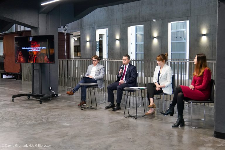 Na krzesłach, obok siebie siedzą cztery osoby. Z lewej strony, na stojaku stoi telewizor
