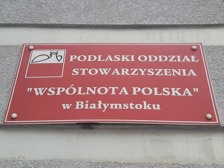Czerwona tabliczka z napisem: Podlaski Oddział Stowarzyszenia "Wspólnota Polska" w Białymstoku