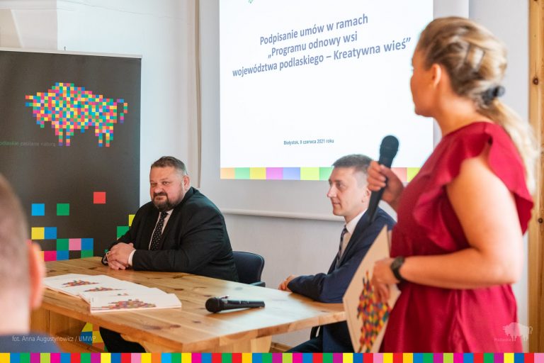 Przy stole konferencyjnym siedzą dwaj mężczyźni, w tle baner z logo woj. podlaskiego