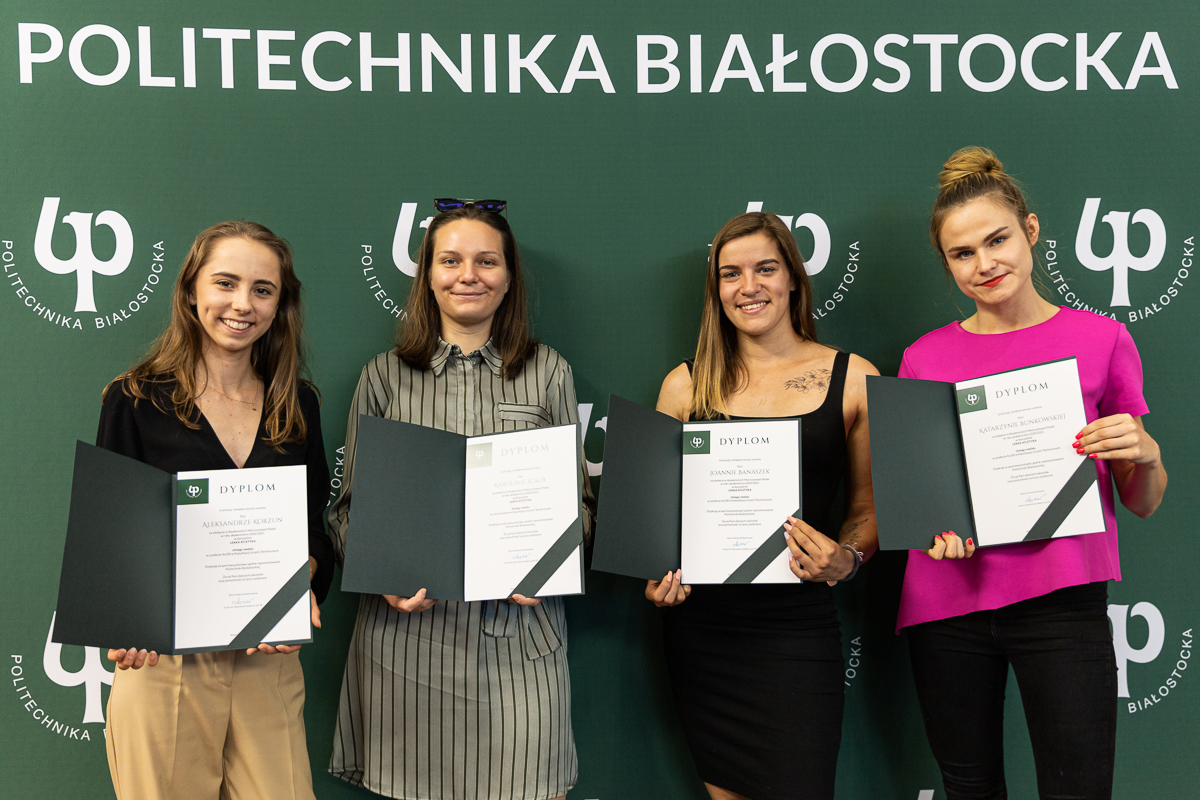Cztery dziewczyny stoją na zielonej ściance Politechniki Białostockiej, przed sobą trzymają dyplomy