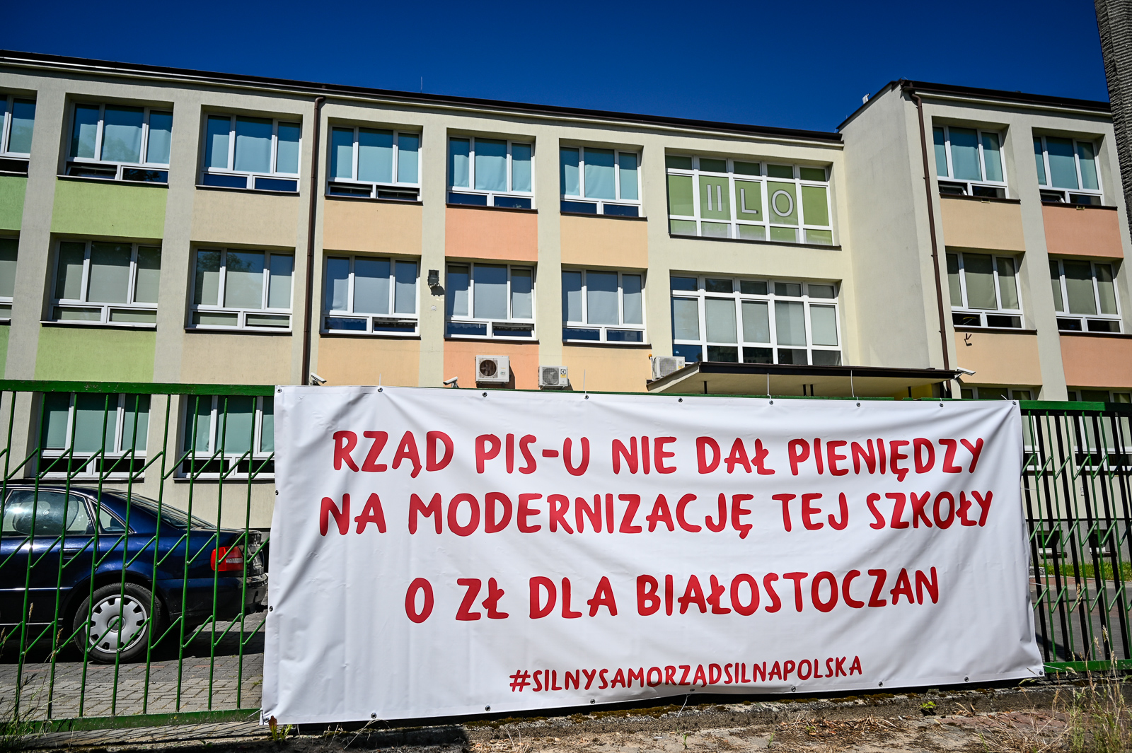 Na ogrodzeniu wisi biały baner z czerwonym napisem: Rząd PIS - u nie dał pieniędzy na modernizację tej szkoły". W tle znajduje się szkoła
