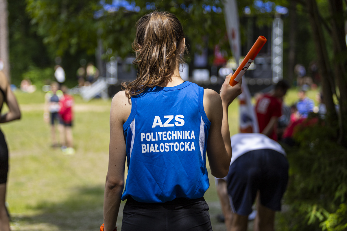 Dziewczyna odwrócona plecami ma na sobie niebieską koszulkę, z białym napisem: AZS Politechniki Białostockiej. W prawej ręce dziewczyna trzyma czerwoną pałeczkę