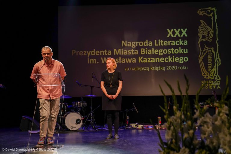 Z lewej strony przy mównicy stoi mężczyzna, który przemawia. Z prawej w tle stoi kobieta, na tle telebimu z napisem XXX Nagroda Literacka Prezydenta Miasta Białegostoku im. Wiesława Kazaneckiego