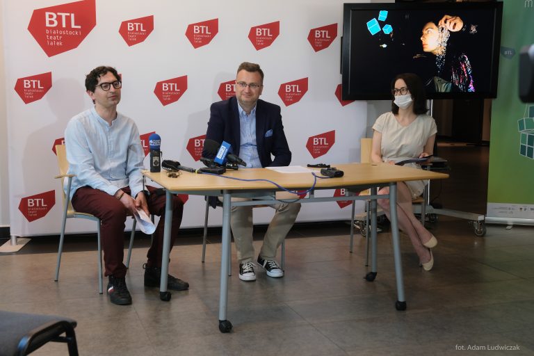 Na tle ścianki z logiem Białostockiego Teatru lalek, przy stole siedzą od lewej dyrektor teatru, zastępca prezydenta Białegostoku oraz pracownica teatru. W górnym prawym rogu znajduje się telewizor, w którym wyświetlany jest fragment spektaklu