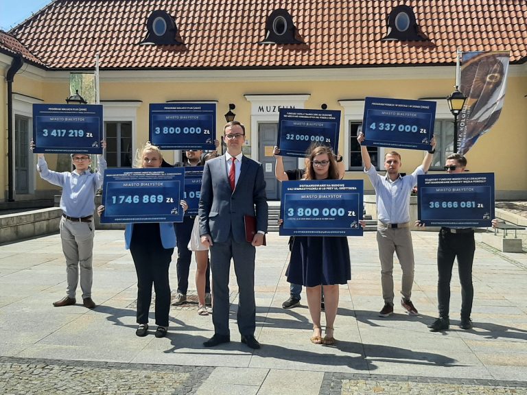 Grupa ludzi stoi na tle Ratusza, w centrum Białegostoku. Osoby te trzymają tabliczki z kwotami dofinansowań, które otrzymał Białystok od rządu