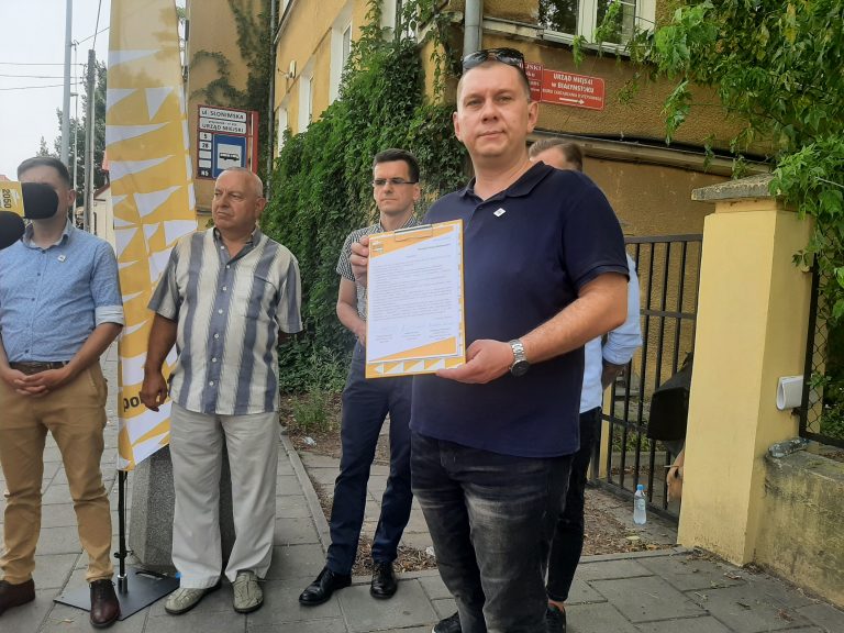 Radny stoi, trzyma w ręku interpelację. Za nim stoją inne osoby
