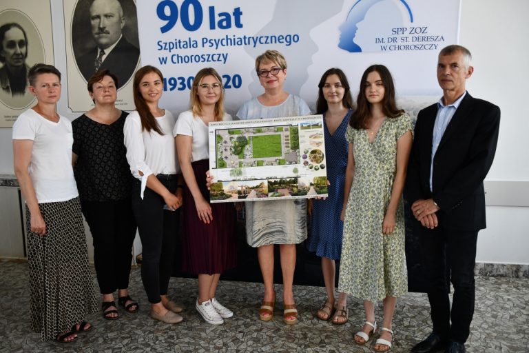 Na ściance z napisem 90. lat Szpitala w Choroszczy stoją m.in. studentki a pośrodku nich dyrektor szpitala, która trzyma w rękach projekt ogrodu przyszpitalnego