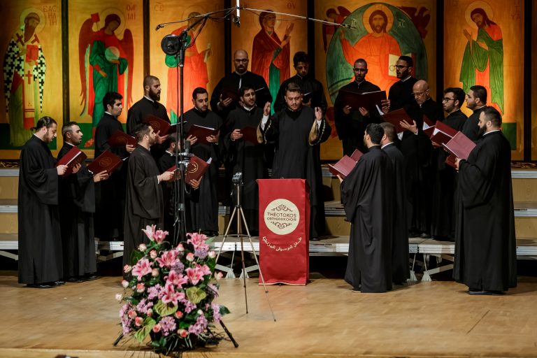 Na tle ikon stoi męski chór prawosławnych księży. Mężczyźni są ubrani w czarne sutanny