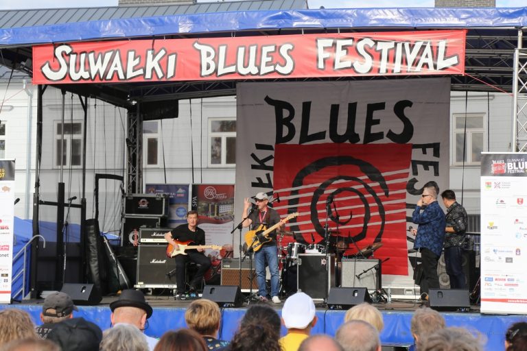 Na scenie, przed którą siedzi publiczność, gra zespół. Nad sceną na czerwonym tle znajduje się biały napis: Suwałki Blues Festival