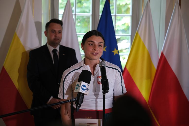 Dziewczyna przemawia do mikrofonu, w tle stoi mężczyzna