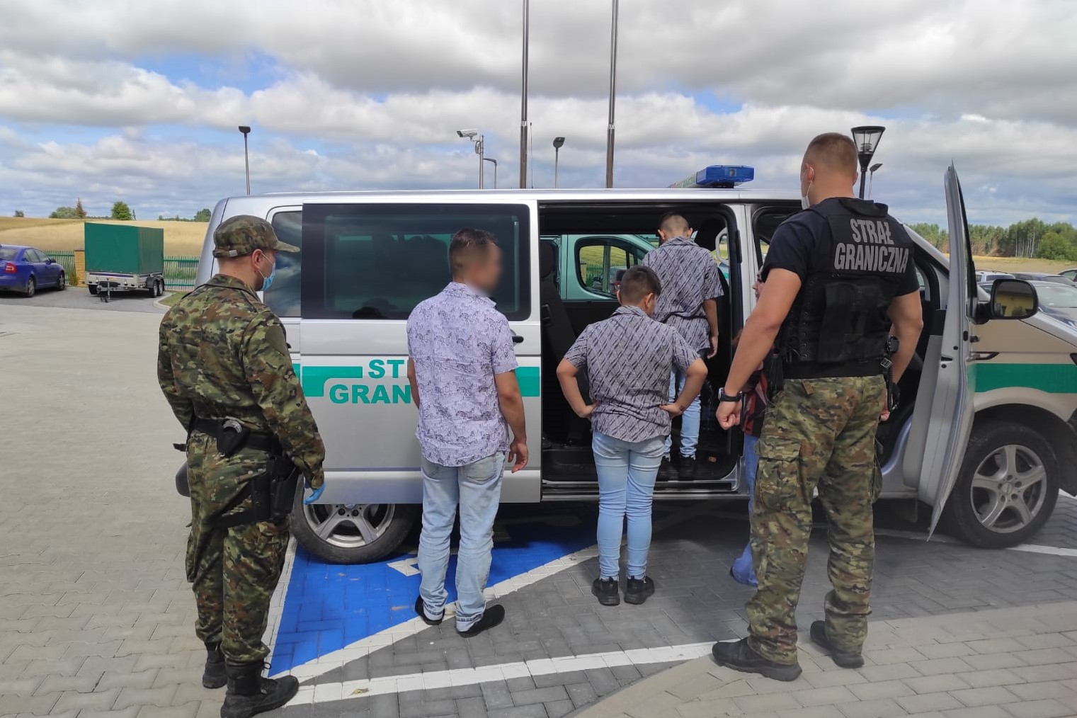 Na parkingu stoją funkcjonariusze POSG, nielegalni migranci wsiadają do służbowego wozu pograniczników