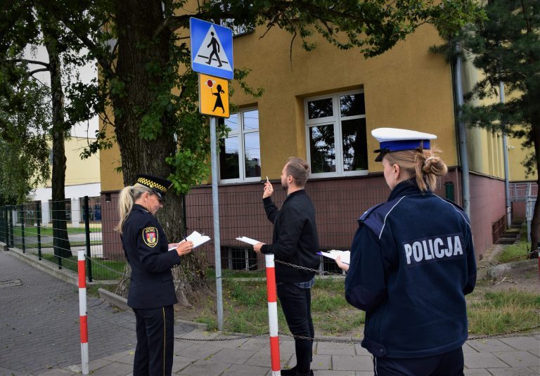 Strażniczka Miejska, Policjantka i urzędnik stoją w okolicy szkoły. Kobiety notują,. mężczyzna przygląda się znakowi drogowemu