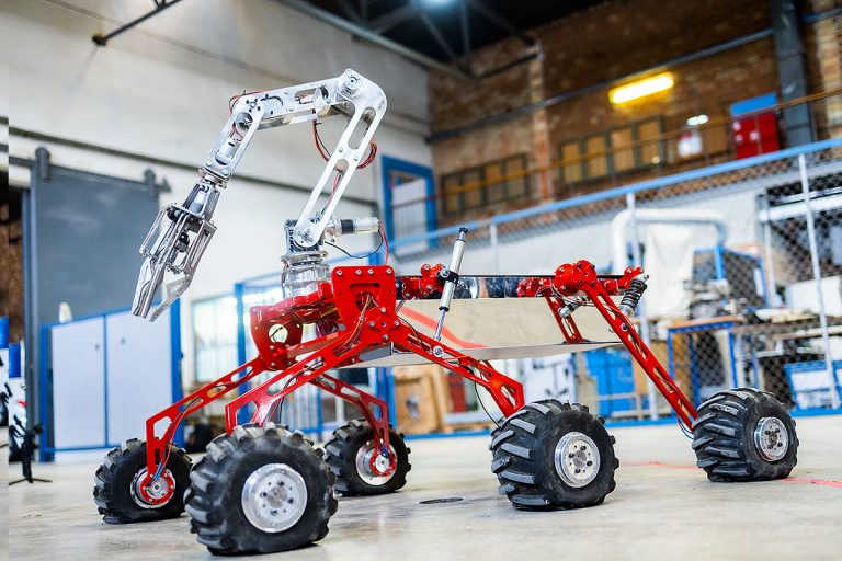 W hali stoi łazik marsjański. Jest sześciokołowym robotem, z białym manipulatorem zakończonym trójpalczastym chwytakiem oraz ma czerwone podwozie
